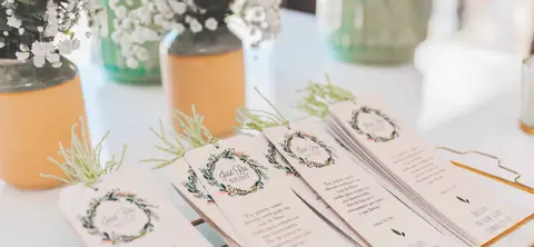 Bílé svatební oznámení na stole s kyticí ve váze v pozadí.